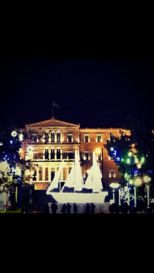 En upplyst byggnad på natten med en glödande segelbåtsinstallation i förgrunden och dekorativa lampor i träden.