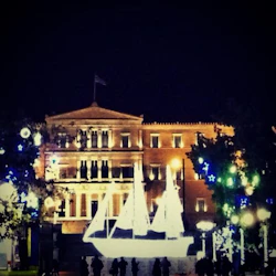 En upplyst byggnad på natten med en glödande segelbåtsinstallation i förgrunden och dekorativa lampor i träden.