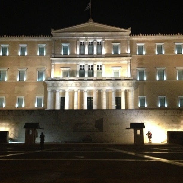 Upplyst neoklassisk byggnad på natten med pelare, fönster och en flagga ovanpå. Vakten står framför nära skuggor.