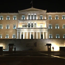 Upplyst neoklassisk byggnad på natten med pelare, fönster och en flagga ovanpå. Vakten står framför nära skuggor.