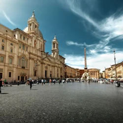 En stor kullerstensplaza med människor som går, med en stor historisk byggnad, en obelisk och en klarblå himmel.
