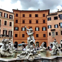 Skulpturfontän som visar Neptunus och mytologiska figurer, på ett torg omgivet av färgglada byggnader med välvda fönster.