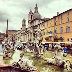 Fontän med utsmyckade statyer på Piazza Navona, Rom. En kupolformad kyrka och gula byggnader syns under en molnig himmel.