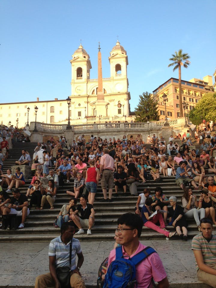 A large crowd of people sitting on steps with a historic church and obelisk in the background under a clear sky.