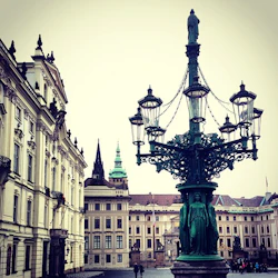 Historiskt torg med utsmyckad gatlampa och klassiska byggnader, inklusive en hög spiral och grön kupol i bakgrunden.