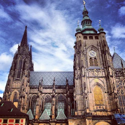 Gotisk katedral med utsmyckade detaljer, stor klocka och spetsiga spiror under en delvis molnig blå himmel.