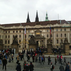 Folkmassa som går på ett kullerstensbelagt torg framför en utsmyckad historisk byggnad med statyer, flaggor och två höga spiror.