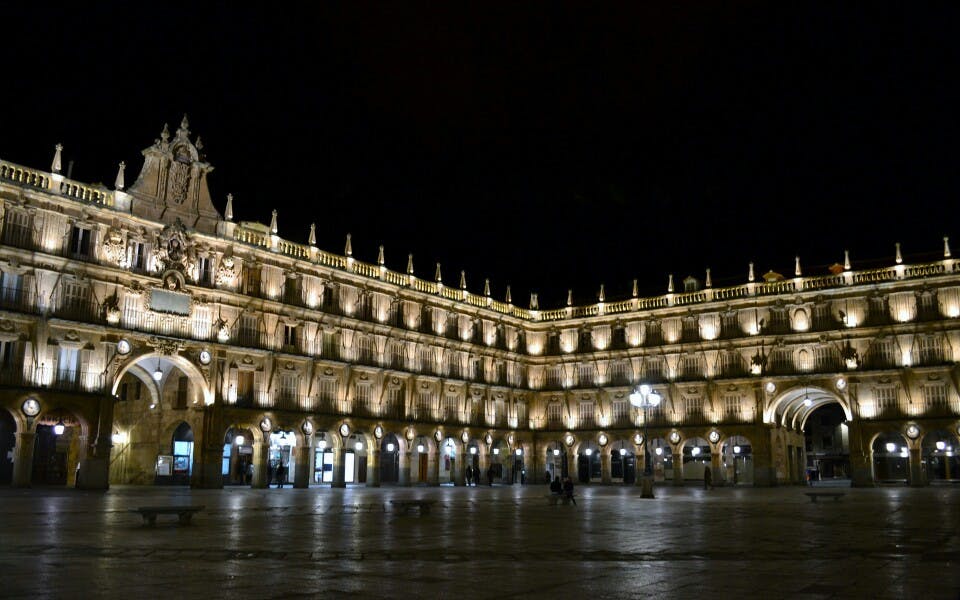 Upplyst historisk byggnad med flera valv och invecklade sniderier på natten, belägen i ett stort öppet torg med få människor.