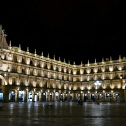 Upplyst historisk byggnad med flera valv och invecklade sniderier på natten, belägen i ett stort öppet torg med få människor.