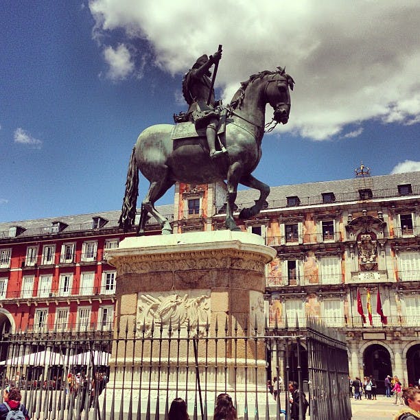 Ryttarstaty i brons på en stenpiedestal på ett torg med en historisk byggnad och flaggor i bakgrunden under en delvis molnig himmel.