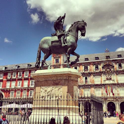 Ryttarstaty i brons på en stenpiedestal på ett torg med en historisk byggnad och flaggor i bakgrunden under en delvis molnig himmel.