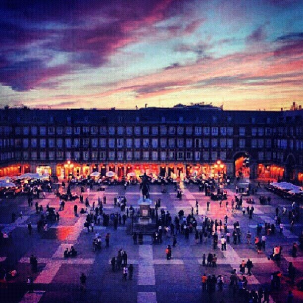 En fullsatt torgscen i skymningen, med människor samlade runt en central staty och byggnader upplysta i bakgrunden under en färgstark himmel.