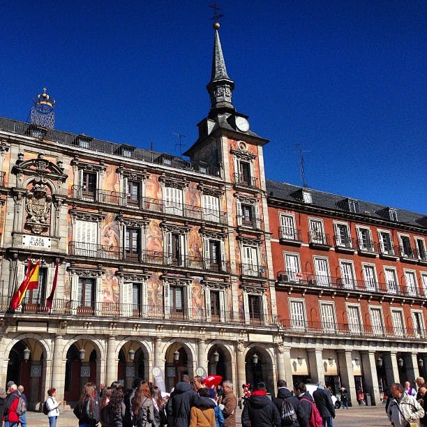 En historisk byggnad med valv, väggmålningar och balkonger under en klarblå himmel. Människor samlas på torget nedanför.