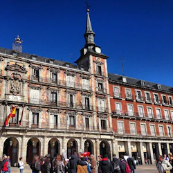 En historisk byggnad med valv, väggmålningar och balkonger under en klarblå himmel. Människor samlas på torget nedanför.