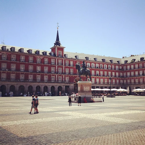 Ett stort torg med en central ryttarstaty, omgivet av en röd byggnad med valv och ett torn. Få människor går runt.