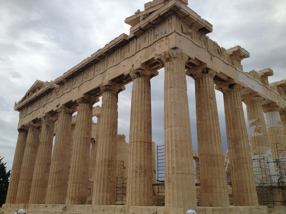 Forntida tempelruiner med höga stenpelare under en molnig himmel, med delvis synliga byggnadsställningar i bakgrunden.