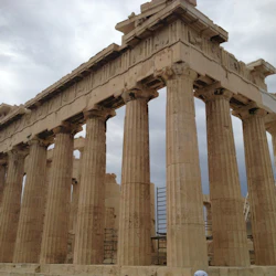 Forntida tempelruiner med höga stenpelare under en molnig himmel, med delvis synliga byggnadsställningar i bakgrunden.