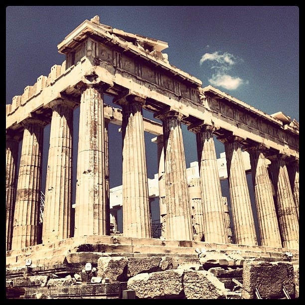 Forntida grekiska tempelruiner med doriska pelare och ett delvis kollapsat tak, under en klar himmel med ett litet moln.