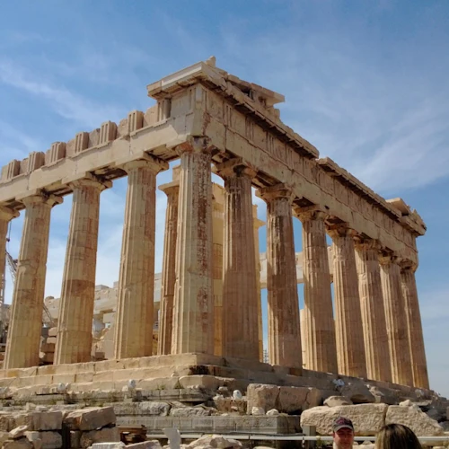Forntida grekiska tempelruiner med höga pelare mot en delvis molnig himmel, med människor synliga i förgrunden.