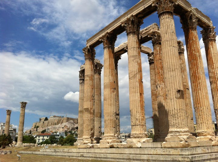 Forntida grekiska tempelruiner med höga, vittrade pelare under en delvis molnig himmel. Avlägsen vy av en sluttande struktur.