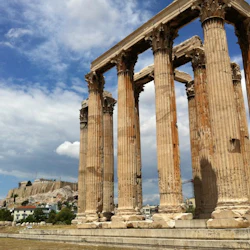 Forntida grekiska tempelruiner med höga, vittrade pelare under en delvis molnig himmel. Avlägsen vy av en sluttande struktur.
