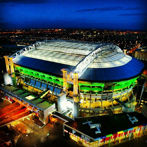 En stor, upplyst stadion med ett infällbart tak i skymningen, omgivet av en stadsbild med blinkande ljus.