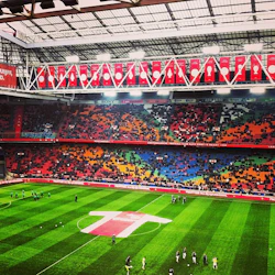 Stadion med färgglada säten, en plan och spelare som värmer upp. Röda banderoller hänger ovanför. Sparsam publik syns på läktaren.