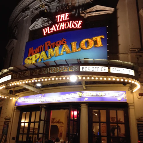 Nattvy av entrén till The Playhouse med starkt upplysta skyltar för "Monty Python's Spamalot.” Folk kommer in.