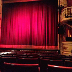 En tom teater med röda säten vetter mot en scen med stängda röda gardiner. Utsmyckade balkonger och skulpturer syns på sidorna.