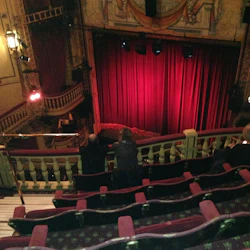 Inuti en teater, röda gardiner på scenen, utarbetad takfresco, svag belysning och några personer som sitter på balkongen.