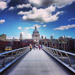 Människor som går på en modern bro mot en historisk kupolformad byggnad under en himmel med spridda moln.
