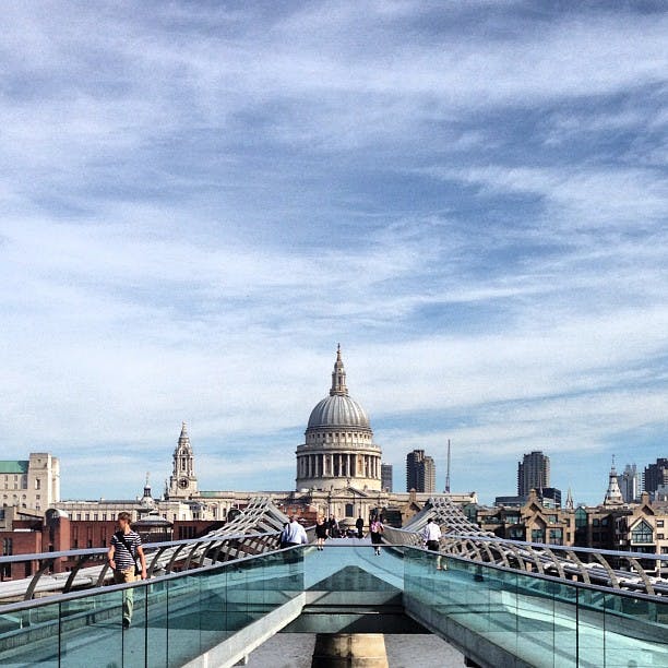People walking on a modern footbridge toward a large dome-shaped building under a partly cloudy sky.