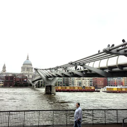 En man står vid floden under en modern gångbro. Stadshus och en kupol syns över vattnet.