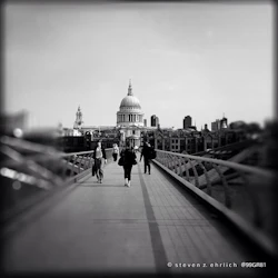Människor går på en bro som leder till en stor kupolbyggnad i en stad, med andra strukturer synliga i bakgrunden.