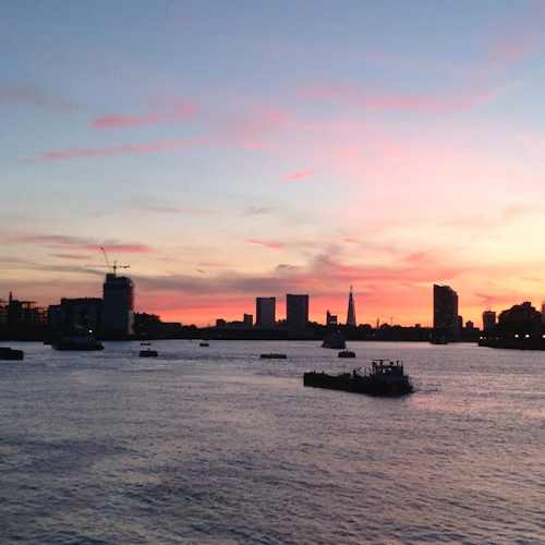 Flod vid solnedgången med utspridda byggnader och båtar silhuett mot en färgglad himmel med rosa och orange nyanser.