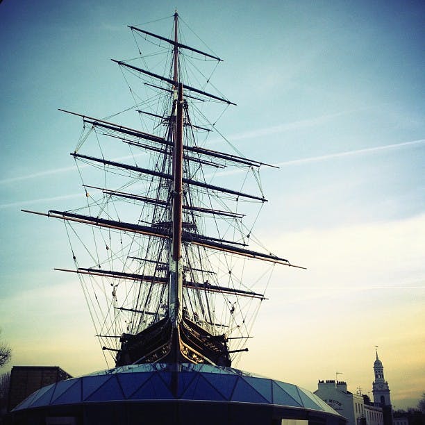 Högt skepp med komplex riggning, dockad ovanpå en modern glaskonstruktion under en blå himmel, med byggnader i bakgrunden.