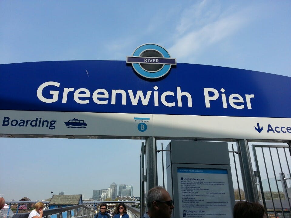 Skylt "Greenwich Pier" med ikoner för ombordstigning och tillgänglighet. Människor och byggnader syns i bakgrunden.