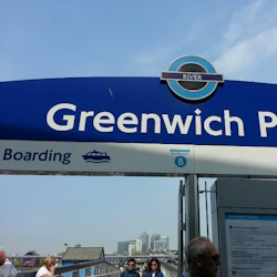 Skylt "Greenwich Pier" med ikoner för ombordstigning och tillgänglighet. Människor och byggnader syns i bakgrunden.