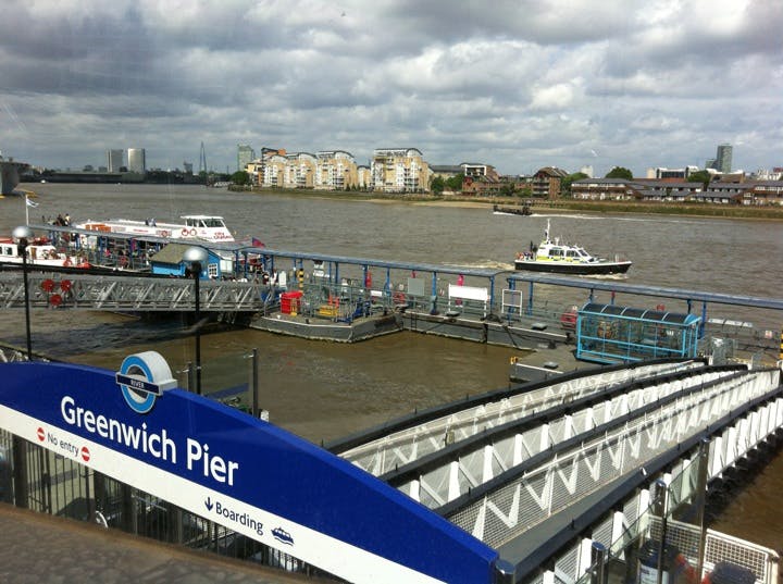 Greenwich Pier med båtar dockade, Thames River och byggnader på motsatta stranden under en molnig himmel.