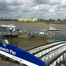Greenwich Pier med båtar dockade, Thames River och byggnader på motsatta stranden under en molnig himmel.