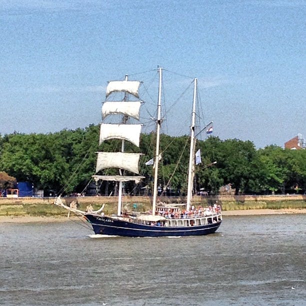 Stor segelbåt med vita segel på en flod med passagerare på däck. Gröna träd och byggnader finns i bakgrunden.