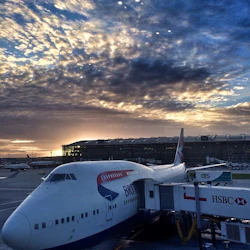 Ett British Airways-flygplan dockade vid en flygplatsport med en terminal och en dramatisk solnedgångshimmel i bakgrunden.