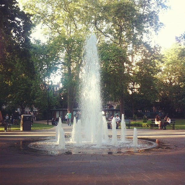 Fontän som sprutar vatten i en park, omgiven av bänkar, människor och höga träd i bakgrunden.