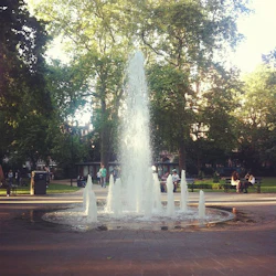 Fontän som sprutar vatten i en park, omgiven av bänkar, människor och höga träd i bakgrunden.