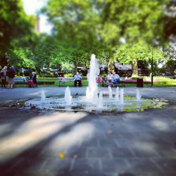 En parkplats med en central fontän, människor som sitter på bänkar och grönskande träd i bakgrunden.