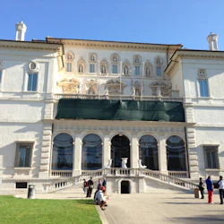 Stor, vit, utsmyckad byggnad med välvda fönster och statyer. Folk sitter och går på stigen framför, med en klarblå himmel ovanför.