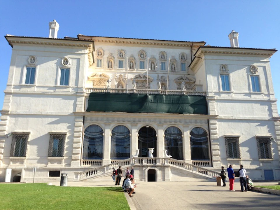 Large, white, ornate building with arched windows and statues. People sit and walk on the pathway in front, with a clear blue sky above.