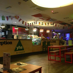 Ett receptionsområde med YHA-märkning, matdisk, flaggor som hänger över huvudet, bänkar, bord, menykort och svag belysning.