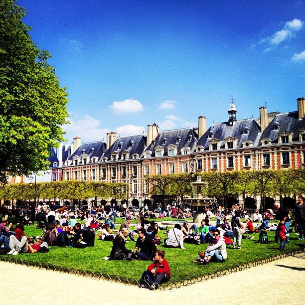 People relax on the grass in a park with a historic building and fountain in the background under a bright blue sky.