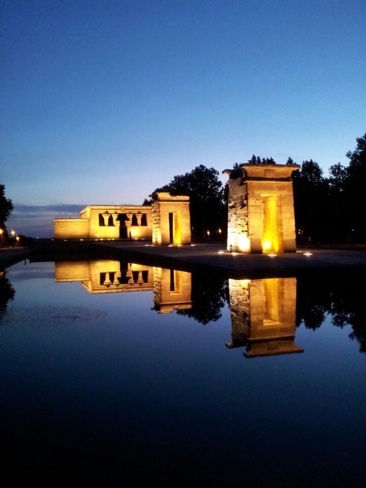 Tända stentempelstrukturer och deras reflektioner i en lugn vattenpool i skymningen, med en klar kvällshimmel och träd i bakgrunden.
